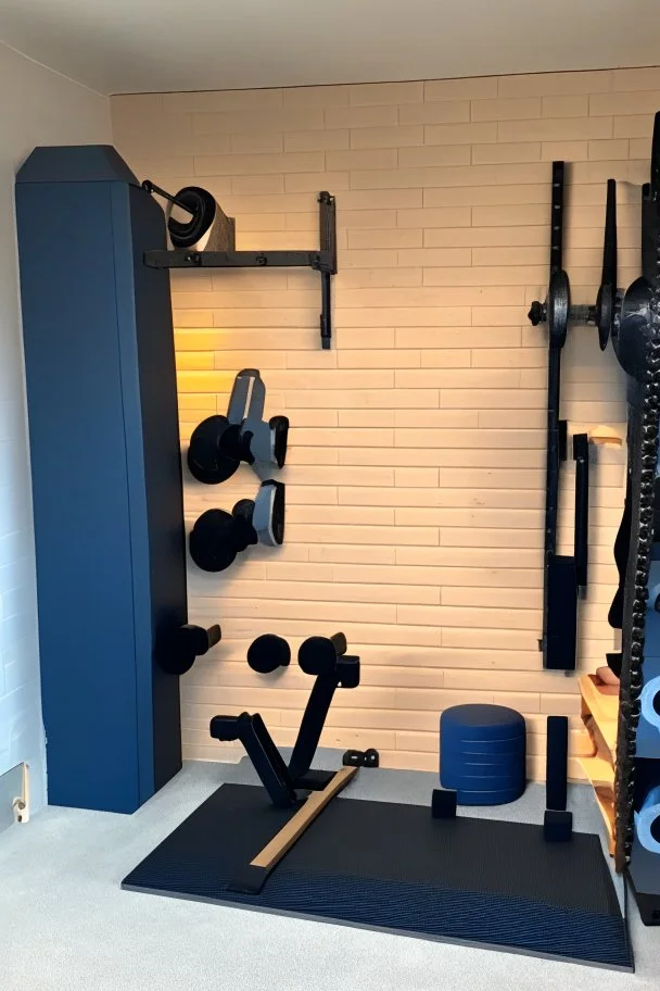 Home gym, wall with a kabinet with weights and small gym equipment. The other side a plyo box and wall balls. In between the plyo box and kabinet a folded weightlifting bench.