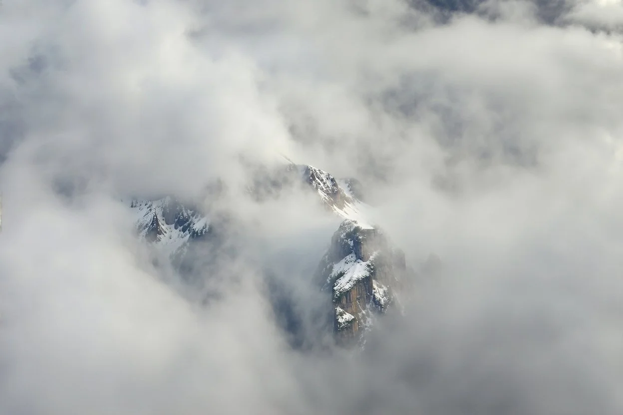 a massive sheer snowy mountain cliff with very sparse vegetation scaling vertically into the sky, partially obscured by dense clouds(color d0d1d5) and mist. the borders — top, bottom, left, and right — fade smoothly into thick fog, while the center reveals the steep, far away rocky cliff face with fine texture and detail. atmospheric lighting, cinematic composition, natural colors, high contrast between fog and stone. photography