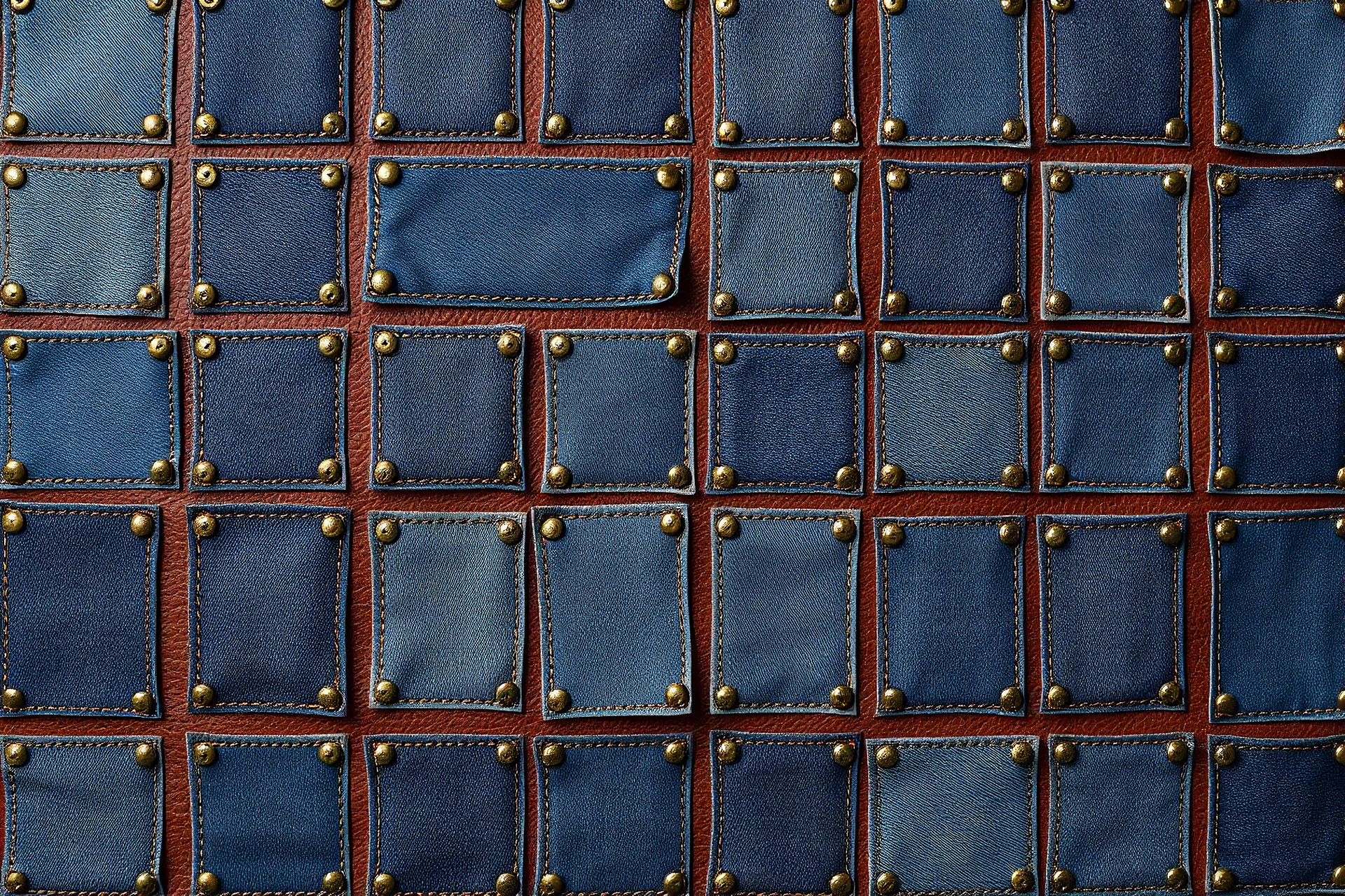 leather background covered with a grid of many rectangular weathered and faded denim patches (navy blue) that are each stitched (brown) and nailed (brass) around the edges