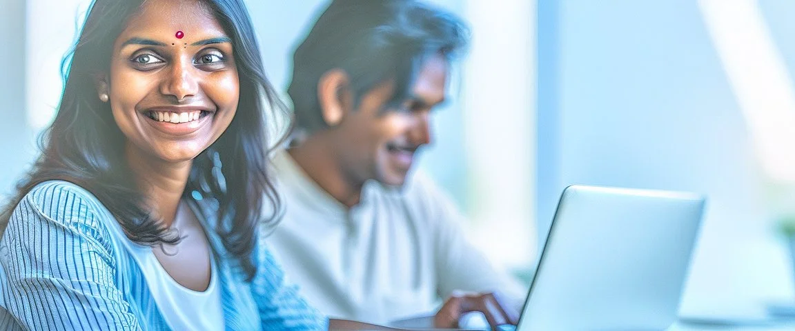 Indian woman sitting at desk with laptop, she looks away from computer and smiles, Indian man blurred in background also sitting at desk working on laptop, business photography