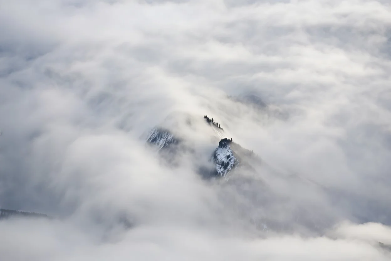 large scale, far away. a massive sheer snowy mountain cliff with very sparse vegetation scaling vertically into the sky, partially obscured by dense clouds(color d0d1d5) and mist. the borders — top, bottom, left, and right — fade smoothly into thick fog, while the center reveals the steep, far away rocky cliff face with fine texture and detail. atmospheric lighting, cinematic composition, natural colors, high contrast between fog and stone. photography