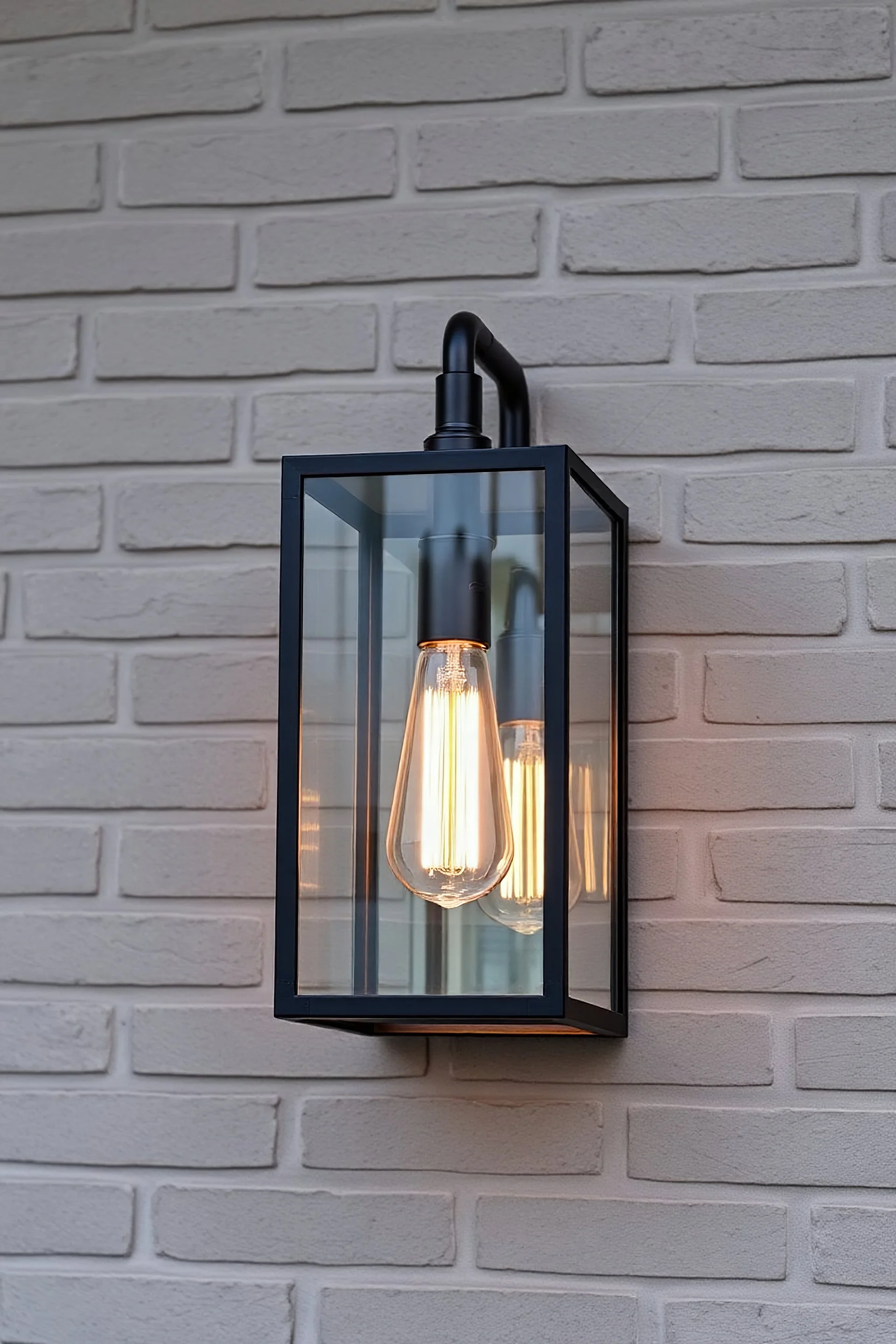 An industrial-style outdoor wall light fixture mounted on a gray brick wall. The light has a rectangular black metal frame with clear glass panels, enclosing a tall filament bulb that emits a warm glow. The texture of the brick wall enhances the modern, minimalist design of the fixture, creating a cozy and inviting ambiance