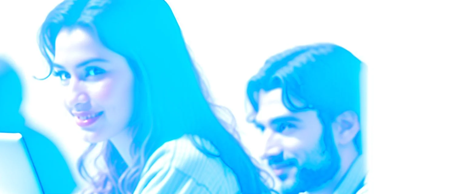 Indian woman sitting at desk with laptop, she looks away from computer and smiles, Indian man blurred in background also sitting at desk working on laptop, business photography