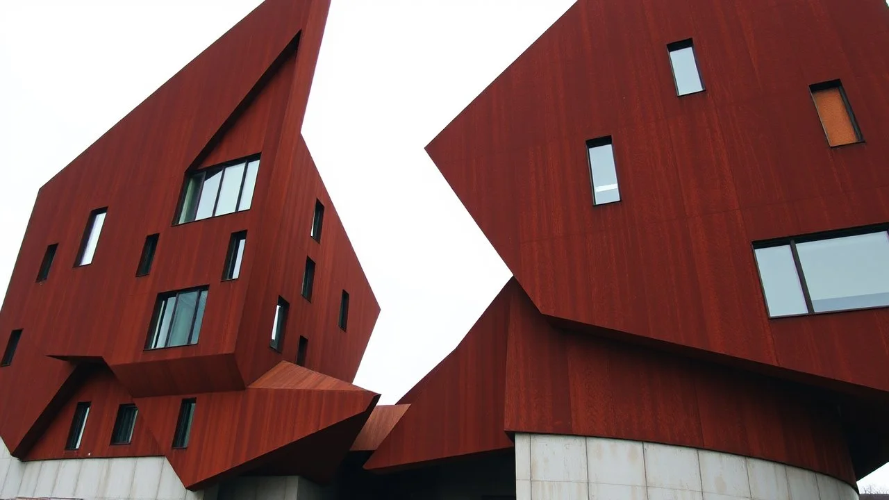 an imposing deconstructivist architectural structure made of weathered corten steel, its surfaces exhibiting a rich patina of rust and streaks. The building is composed of sharp, angular geometric forms that jut out at various angles, creating a dynamic and almost chaotic silhouette against a pale, overcast sky. Numerous rectangular windows, some tinted a pale blue or green, are embedded within the metal planes, offering glimpses into the interior. The structure rests on a base of light-colored