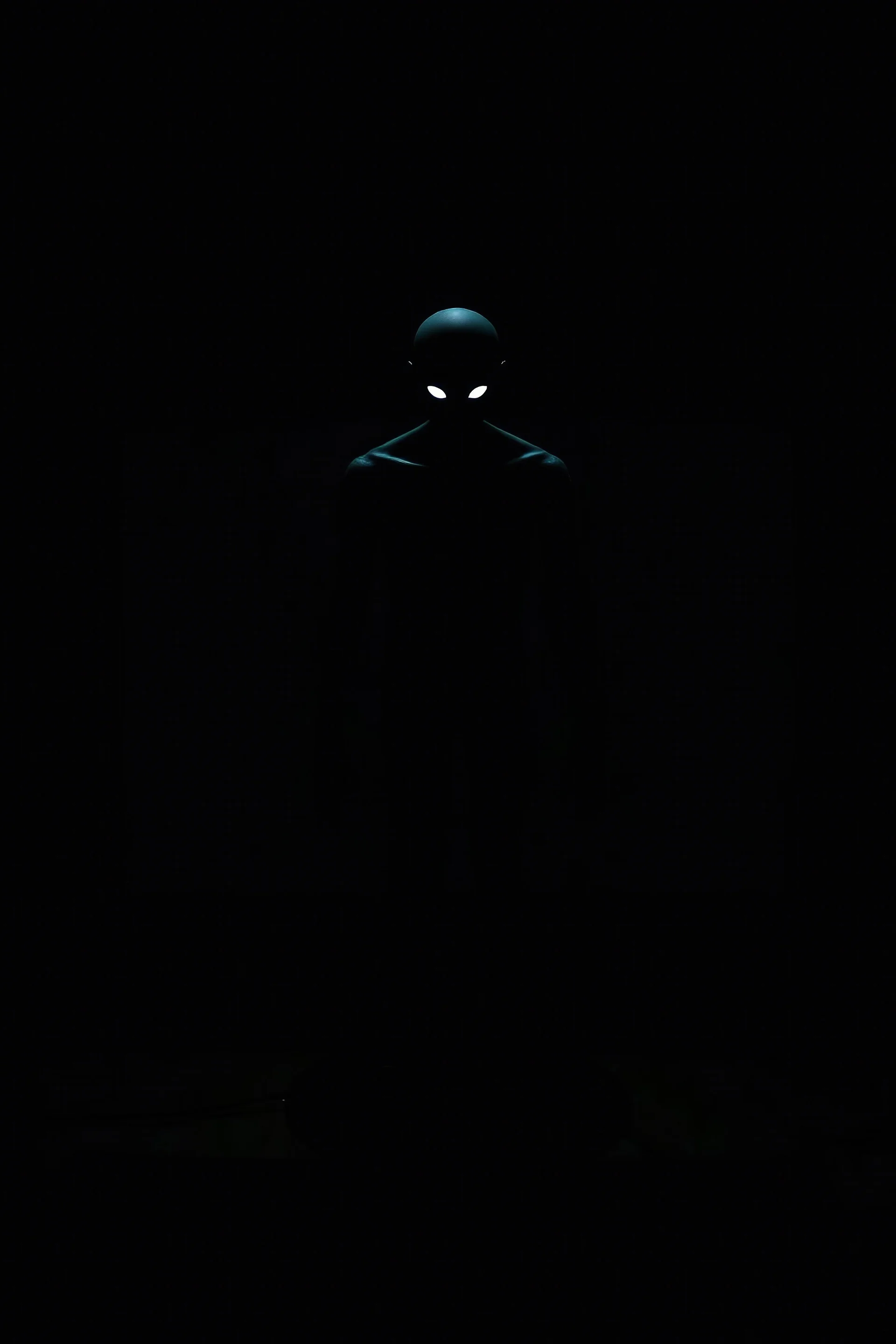 Inside a dark, shadow-filled computer screen, a sinister humanoid silhouette emerges. The silhouette has a tall, gaunt figure with elongated limbs and a bald head. Its hollow black eyes glow faintly, directly fixating on the viewer, creating an intense and unnerving connection. The monstrous figure is centrally positioned within the screen, enhancing the sense of dread and malevolent presence emanating from within the digital realm. The surrounding room remains dark and oppressive, with minimal