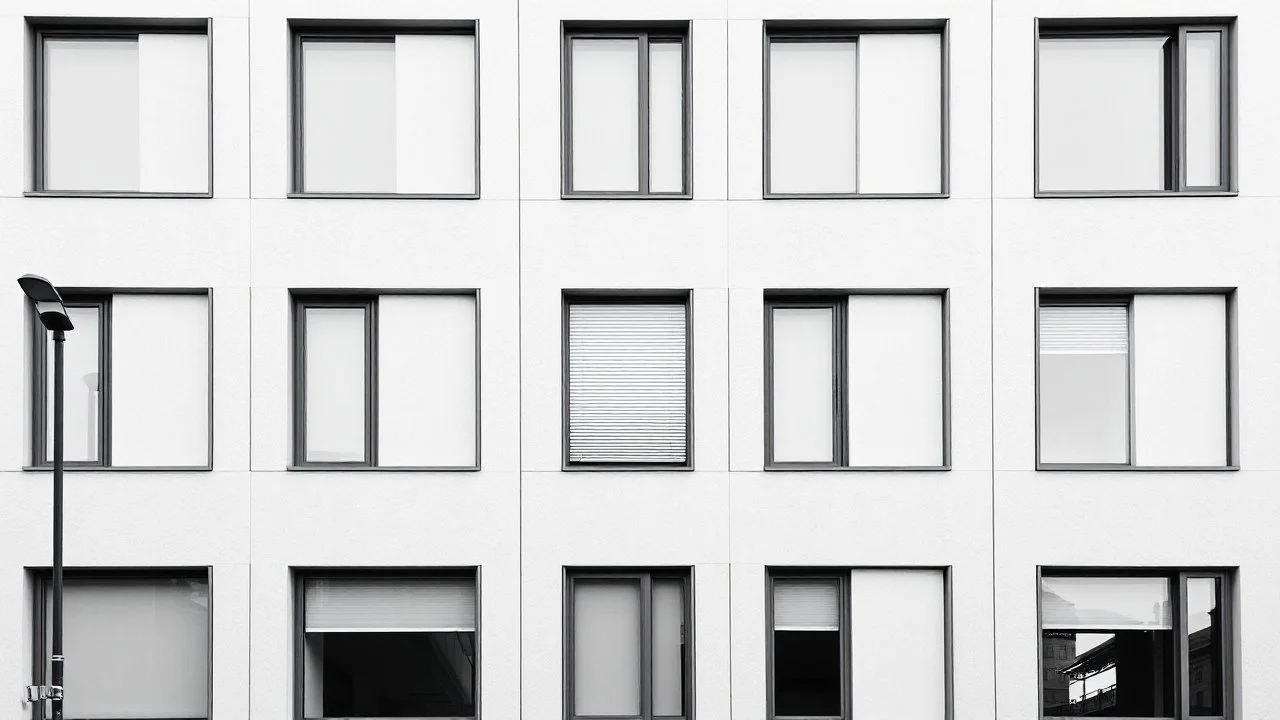 Image is a black and white photograph of a modern building facade. The layout features a symmetrical arrangement of large, rectangular windows with sleek, minimalistic frames. The windows vary in size and are positioned at different heights, creating a dynamic visual pattern. The building's surface is smooth and unadorned, with a slightly textured finish. A single street lamp is visible on the left side of the image, adding an element of urban infrastructure. The overall style is contemporary, w