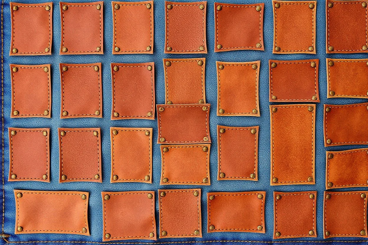 blue denim background covered with a grid of many rectangular weathered brown leather patches that are each stitched around the edges