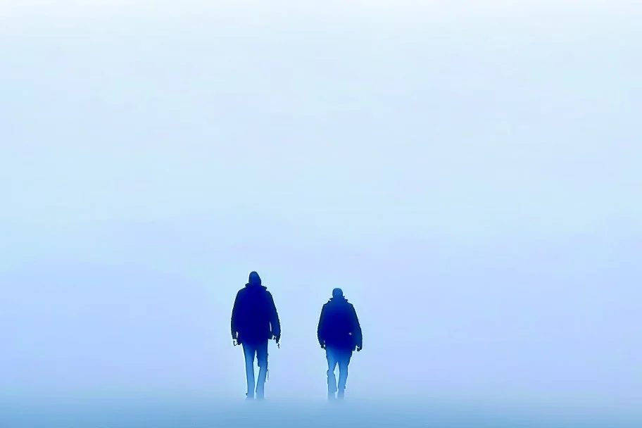 two people seen from behind walking side by side in an empty foggy plain, above there is blue sky