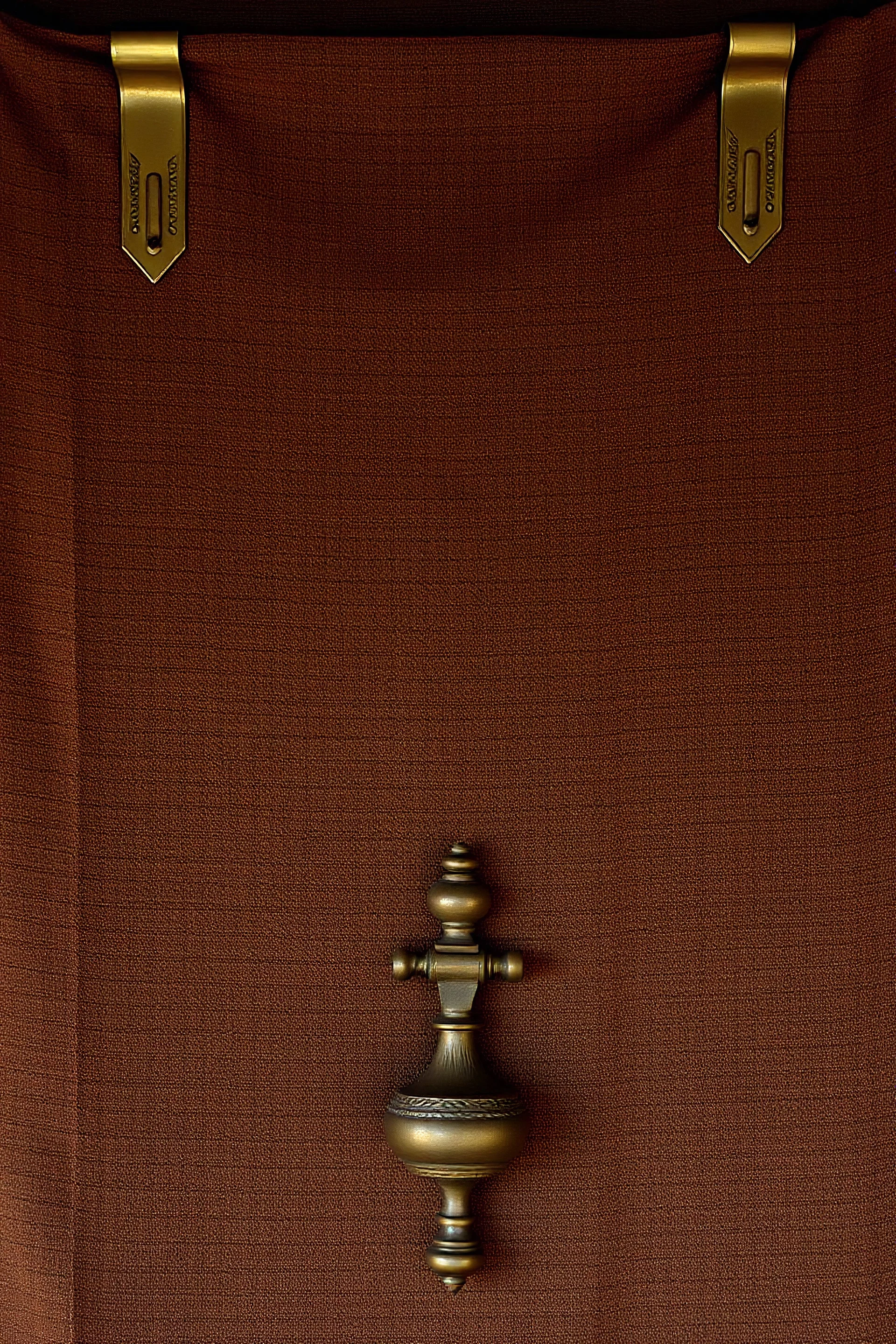 darker textured hanging fabric background, with decorative brass corners at top, and a heavy brass decorative element attach across the bottom