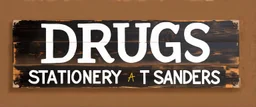 a hand painted sign on dark painted wood planks, first line says "DRUGS", second line (Smaller) says "STATIONERY T SANDERS", on a brown background
