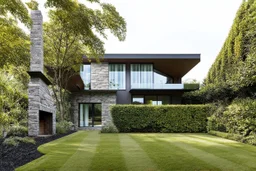 Modern house in the middle of lush vegetation. It is a mix of stone and glass, with a prominent fireplace to the left. Large windows and glass doors. It has several levels, with a balcony on the ground floor. The surrounding area is adorned with tall trees, bushes and manicured lawns. The general atmosphere is serene, in a quiet residential area, architecture, vibrant