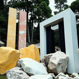 Un'immagine di un paesaggio naturale con marmi gialli e rossi pastello imponenti accanto a strutture artificiali verticali e alberi. Queste strutture sembrano blocchi rettangolari riflettenti, come colonne di vetro, che creano un contrasto tra il naturale e l'artificiale. Gabbia brutalista si erge solitaria. Persona osserva. bianco con venature e solchi. Tenda nera scende a terra. La scena trasmette un senso di sospensione e dialogo tra elementi organici. Dettagli delle rocce molto accentuati