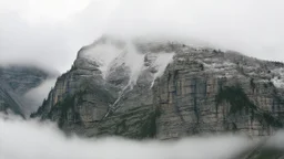 a massive sheer snowy mountain cliff with very sparse vegetationscaling vertically into the sky, partially obscured by dense clouds and mist. the borders — top, bottom, left, and right — fade smoothly into thick fog, while the center reveals the steep, rocky cliff face with fine texture and detail. atmospheric lighting, cinematic composition, natural colors, high contrast between fog and stone.