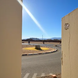 Fotografia di contesto esterno, una parte di un edificio. A destra c'è un muro con della vernice scrostata e su di esso è appeso un fascio di luce. Deserto. Lo sfondo mostra un vialetto asfaltato e una piccola area gialla con una roccia alcune case in lontananza. Persone. Il cielo è blu. L'immagine trasmette una sensazione di tranquillità e semplicità quotidiana.