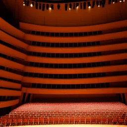 a single chair on stage in the beam of single spotlight facing empty audience at a dark and empty symphony hall