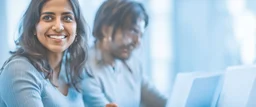 Indian woman sitting at desk with laptop, she looks away from computer and smiles, Indian man in background also sitting at desk working on laptop but blurred, business photography
