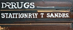 a hand painted sign on dark painted wood planks, first line says "DRUGS", second line (Smaller) says "STATIONERY T SANDERS", on a brown background