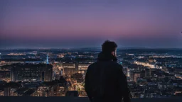 An Englishman in a bomber jacket standing to one side of a tall building looking across a city after sunset