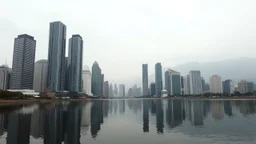 A cityscape of tall, modern skyscrapers reflected in a calm body of water, with mountains in the background under a hazy, overcast sky