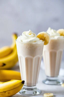 A detailed close-up of banana milkshakes, captured in a vibrant and appetizing food photograph. The milkshakes are presented in tall glasses, topped with whipped cream and garnished with fresh banana slices. The background is blurred to focus on the milkshakes, with soft, natural lighting that highlights the creamy texture and burst of flavor. The image evokes a sense of indulgence and delight, making the viewer crave a sip.