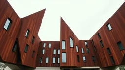 an imposing deconstructivist architectural structure made of weathered corten steel, its surfaces exhibiting a rich patina of rust and streaks. The building is composed of sharp, angular geometric forms that jut out at various angles, creating a dynamic and almost chaotic silhouette against a pale, overcast sky. Numerous rectangular windows, some tinted a pale blue or green, are embedded within the metal planes, offering glimpses into the interior. The structure rests on a base of light-colored