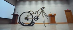 pinhole cam, airbrush 3d rendering, on tv screen:A five-second cinematic single take in a clean industrial presentation hall. A bizarre mechanically plausible tracked bicycle machine stands beside a presentation podium, designed to look like a giant mechanical praying mantis. Two large bicycle wheels form its main body and four thin articulated mechanical legs support it. Two long folded mechanical front arms resemble mantis forelegs