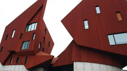an imposing deconstructivist architectural structure made of weathered corten steel, its surfaces exhibiting a rich patina of rust and streaks. The building is composed of sharp, angular geometric forms that jut out at various angles, creating a dynamic and almost chaotic silhouette against a pale, overcast sky. Numerous rectangular windows, some tinted a pale blue or green, are embedded within the metal planes, offering glimpses into the interior. The structure rests on a base of light-colored