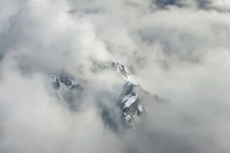 large scale, far away. a massive sheer snowy mountain cliff with very sparse vegetation scaling vertically into the sky, partially obscured by dense clouds(color d0d1d5) and mist. the borders — top, bottom, left, and right — fade smoothly into thick fog, while the center reveals the steep, far away rocky cliff face with fine texture and detail. atmospheric lighting, cinematic composition, natural colors, high contrast between fog and stone. photography