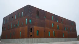 an imposing deconstructivist architectural structure made of weathered corten steel, its surfaces exhibiting a rich patina of rust and streaks. The building is composed of sharp, angular geometric forms that jut out at various angles, creating a dynamic and almost chaotic silhouette against a pale, overcast sky. Numerous rectangular windows, some tinted a pale blue or green, are embedded within the metal planes, offering glimpses into the interior. The structure rests on a base of light-colored