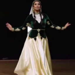 A graceful Azerbaijani female traditional dancer performing a famous Azerbaijani folk dance. She is wearing a beautiful traditional Azerbaijani dance costume: a long flowing silk dress with rich embroidery, wide skirt, and a colorful kelaghayi (traditional Azerbaijani silk headscarf) covering her head. The dancer moves elegantly with soft hand gestures, delicate finger movements, and smooth spinning steps typical of Azerbaijani women’s dances. The atmosphere is festive and cultural, with traditi