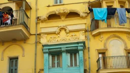 An old, yellow-colored building with ornate architectural details and balconies. There are two people standing on the balcony, and clothes are hanging out to dry. The background suggests an urban, Mediterranean-style setting