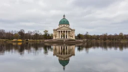 A classical-style building with a green dome stands on a platform extending into calm water. The building and surrounding trees are reflected in the water under a cloudy sky.