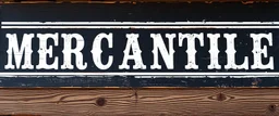 fill composition with large and wide hand painted old western sign that reads: "MERCANTILE" white on black with inset white stripe aged paint on wood sanserif