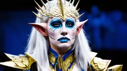The image features a person with an elaborate makeup and costume that gives them an otherworldly appearance. They have pale skin, long white hair, and pointed ears. Their facial features are accentuated by gold and blue makeup, which includes intricate designs around their eyes and on their forehead. The individual is wearing a headdress with a spike-like design that adds to the fantastical look. They are also adorned in a costume with gold and blue elements, featuring what appears to be scales