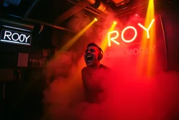 nedsliten sunkig techno club, mycket scen rök i lokalen, och gula röda strobes, en Neon Skylt med texten "DJ ROY" på väggen, DJ Roy skriker rätt in i kameran, och headbangar, euforisk