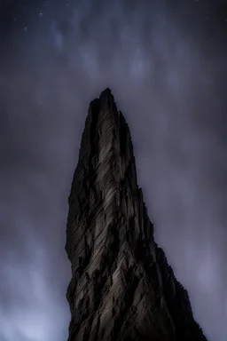 looking up from below at one single mountain pillar cliff sharp face rises out of the misty foreground into the night sky background.