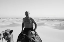 man looking like Walter White in breaking bad, bald, with bare chest riding on a camel in sahara in desert, panorama, black and white photo, staring at a small town in horizon