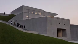 Image showcases a modern architectural structure with a minimalist design, featuring a multi-level building constructed from grey stone. The building has a series of rectangular columns and large windows, each adorned with circular light fixtures. The structure is built into a grassy hillside, with a staircase on the left side leading up to the entrance. The lower level includes a large, grid-patterned door. Several people, of various ages and ethnicities, are walking along the staircase, around