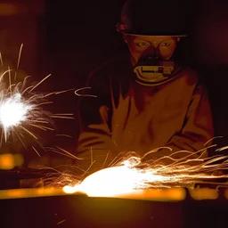 Imagen realista y dramática de una trabajador en una fábrica de fundición rodeado de las chispas que saltan, en medio de la penumbra, primer plano del trabajador mirando la pieza que funde. Fotografía periodística realizada con una cámara Leica y un objetivo de 50 mm