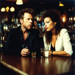 A color photograph of singer Tom Waits sitting at a table in a bar having a drink with a beautiful young woman