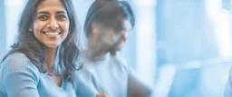 Indian woman sitting at desk with laptop, she looks away from computer and smiles, Indian man blurred in background also sitting at desk working on laptop, business photography