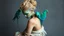 Placeholder: A mystical, ethereal portrait of a woman with luminous green skin, adorned with four vibrant teal parrots perched on her shoulders and back. Her hair is styled in a complex, blonde updo. She is seated, with her head bowed in a pensive posture, her body wrapped in a flowing, textured garment that resembles delicate, organic fibers or scales. The skin has a slightly mottled, almost reptilian texture with hints of coppery undertones, suggesting a connection to nature or otherworldly origins. Her bo