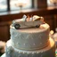 Placeholder: A hyper-realistic, detailed photo of a massive, elegant single-tier wedding cake. On top of the cake, there is a luxurious vintage open-top convertible toy car, made of fondant or edible sugar. Inside the car, sitting happily, are a tiny bride and groom doll. The cake is beautifully decorated with intricate white and silver icing, sugar pearls, and delicate floral patterns. The scene is lit with soft, warm studio lighting, creating a magical and joyful atmosphere. Shallow depth of field, focusin