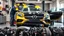 Placeholder: A car repair shop where two people in work clothes are repairing a black and yellow Benz CLS 550 with an eye-catching theme in a sophisticated and special repair shop where at the bottom of the image a collection of car engine parts is arranged on the floor