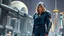 Placeholder: A full-body photograph of a young, slender woman with blond shoulder-length bob hair in a blue jumpsuit, standing in a futuristic cityscape with advanced spacecraft behind them, with a huge android in a huge metal exosuit with a helmet standing guard beside her.