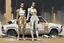 Placeholder: ((3)) determined cyberpunk women (wearing white khiton:2) standing in front of a car wreck, acrylic painting, minimal art, centered, wild sparse brushstrokes, amazing verticals, great parallels, low bleak colors of gold, beige and black, excellent negative space contrasts
