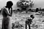 Placeholder: anguished mother and child war refugees, as child stands amidst rubble and ruins with nuclear mushroom cloud in the background. Mother crying in agony. human skulls strewn about. single red flower growing near child.