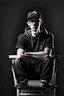 Placeholder: Digital art for a young white man with a eyesglasses and black new balance cap in photo setion sits on a chair and talks to the camera