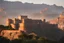 Placeholder: 'Agra fort style', grey granite fortress situated in hilly Highlands seen from far away built into the shadows of granite cliffs with the sunset behind it