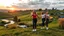 Placeholder: a group of young ladies in sports pants and blouse are dancing to camera in village over high grassy hills,a small fall and river and wild flowers at river sides, trees houses ,next to Ripe wheat ready for harvest farm,cloudy sun set sky